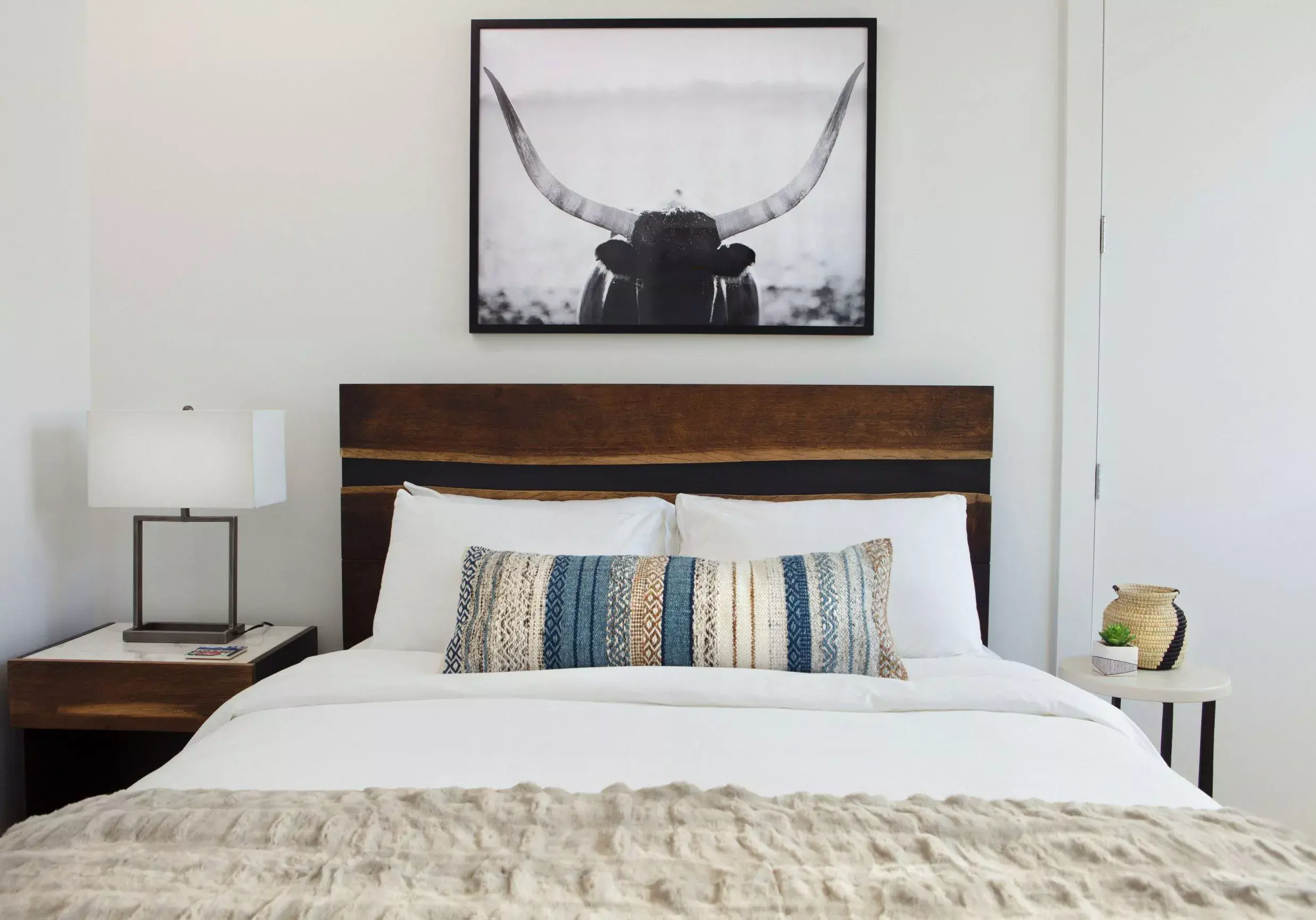 Etch Interior Design A bed made up in white with a wood headboard adorned with one single long lumbar pillow made with cream, beige, and blue colors in textured folds.