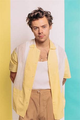 Harry Styles wearing a yellow and white short-sleeve shirt over a white tank top with tan trousers, standing against a pastel color-blocked backdrop.