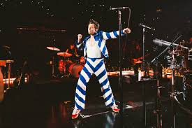 Harry Styles performing on stage in a bold blue and white striped suit with red shoes, arms raised mid-dance.