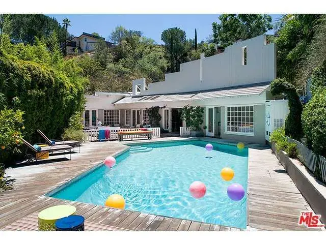 Backyard pool surrounded by a wood deck with colorful beach balls, lounge chairs, and a white modern-style home.