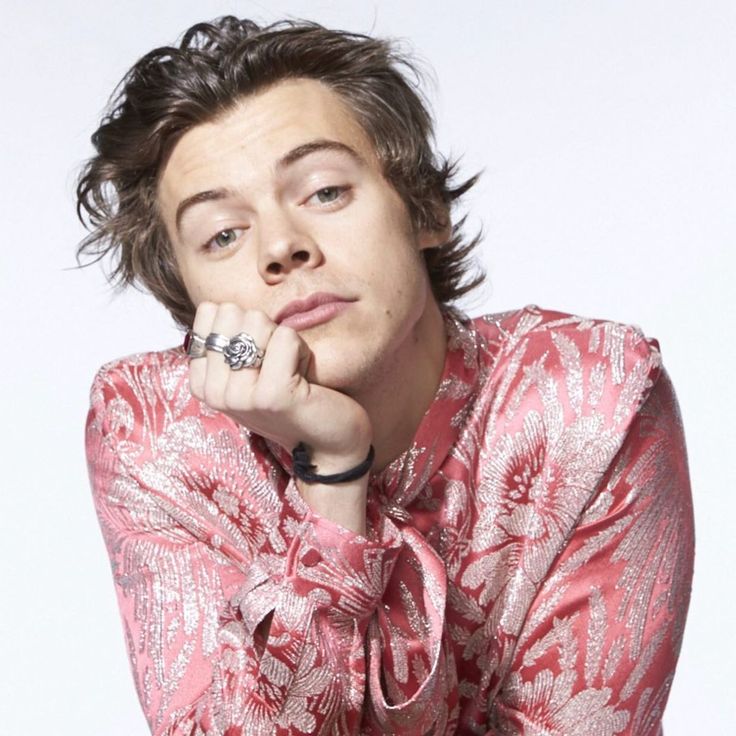 Harry Styles wearing a pink and silver floral satin blouse, resting his chin on his hand with silver rings and tousled hair.