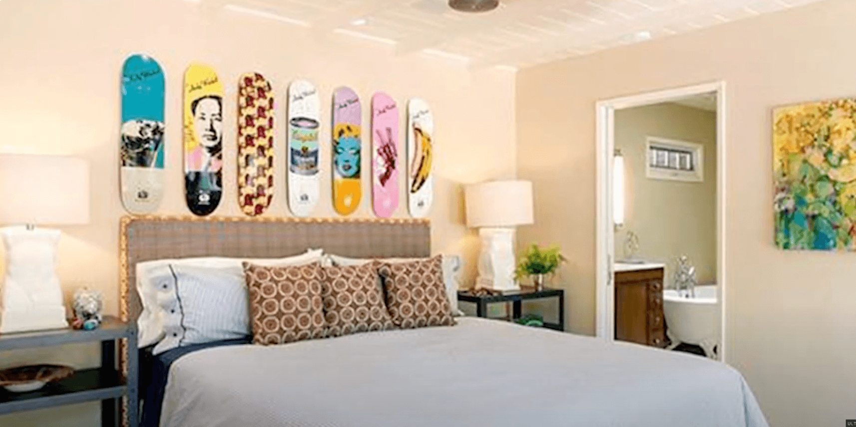 Bedroom with skateboard art above the bed, geometric pillows, white lamps, and a view into a bathroom with a clawfoot tub.
