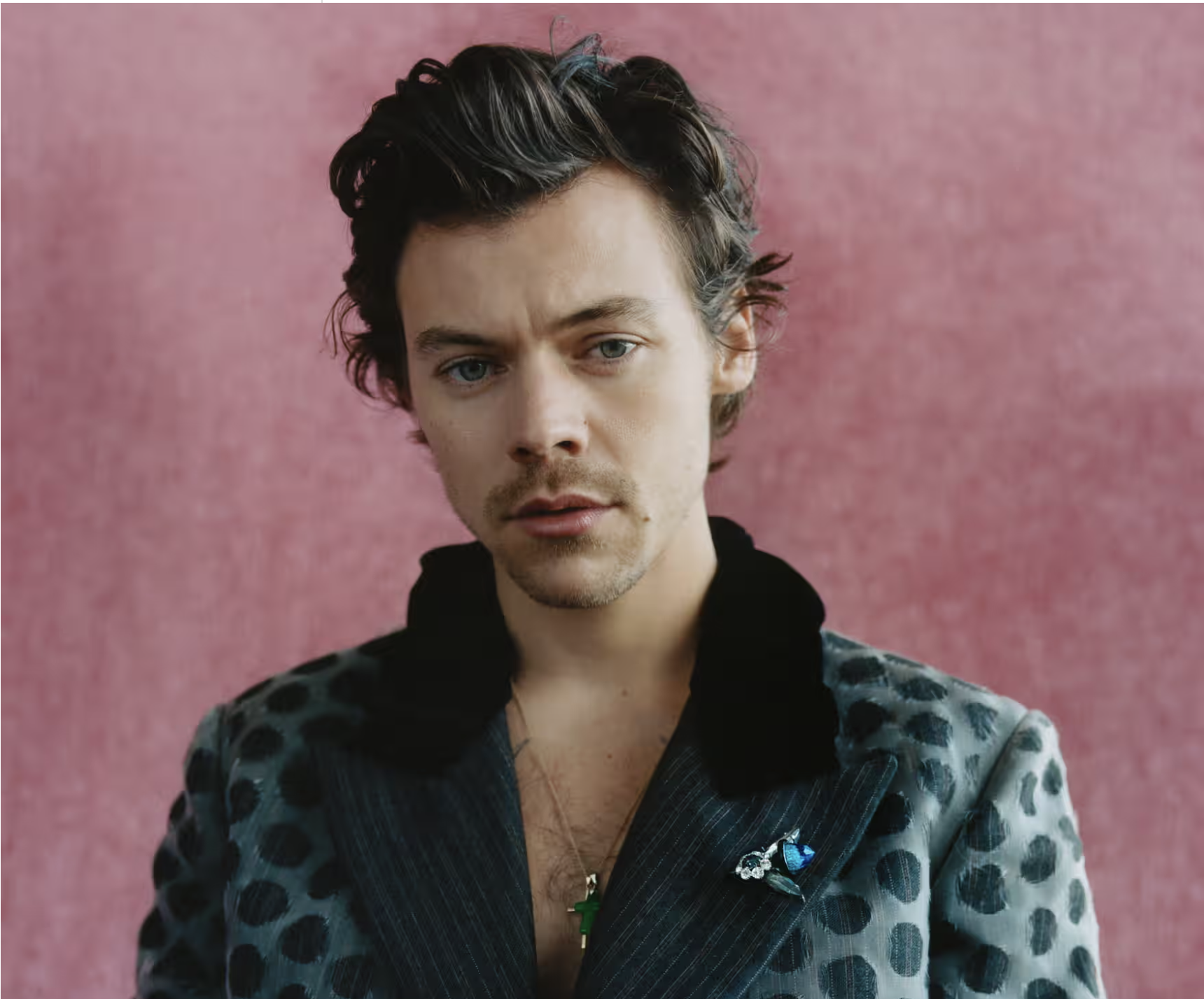 Harry Styles in a teal polka-dot blazer with a velvet collar and blue brooch, standing against a pink textured background in a portrait by Samuel Bradley.