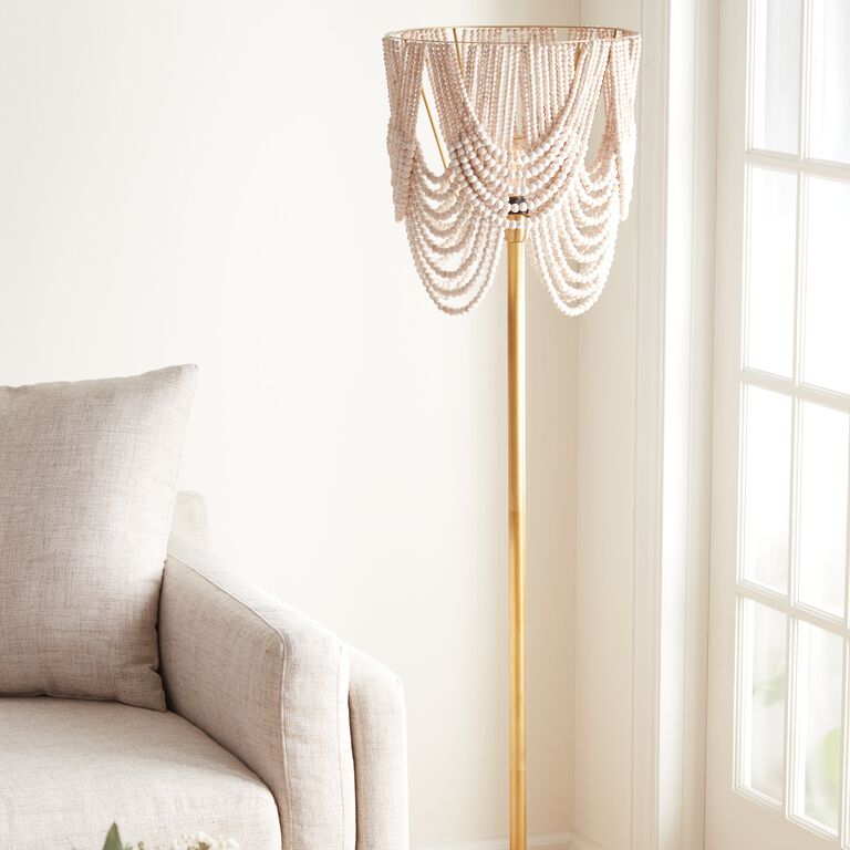 Gold floor lamp with a white beaded draped shade, next to a beige linen sofa in a sunlit room.