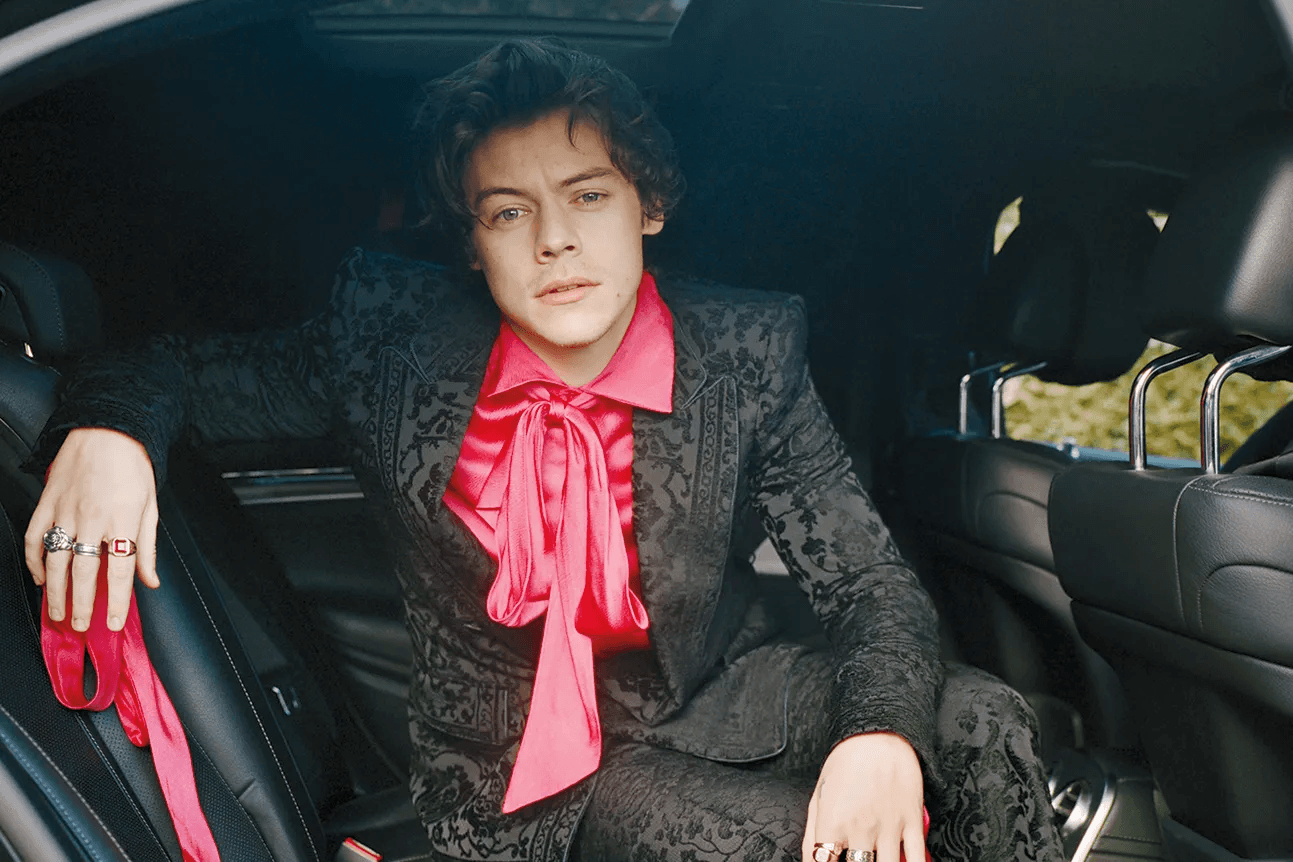 Harry Styles wearing a black damask suit with a hot pink satin bow blouse, seated in a car with rings on both hands.