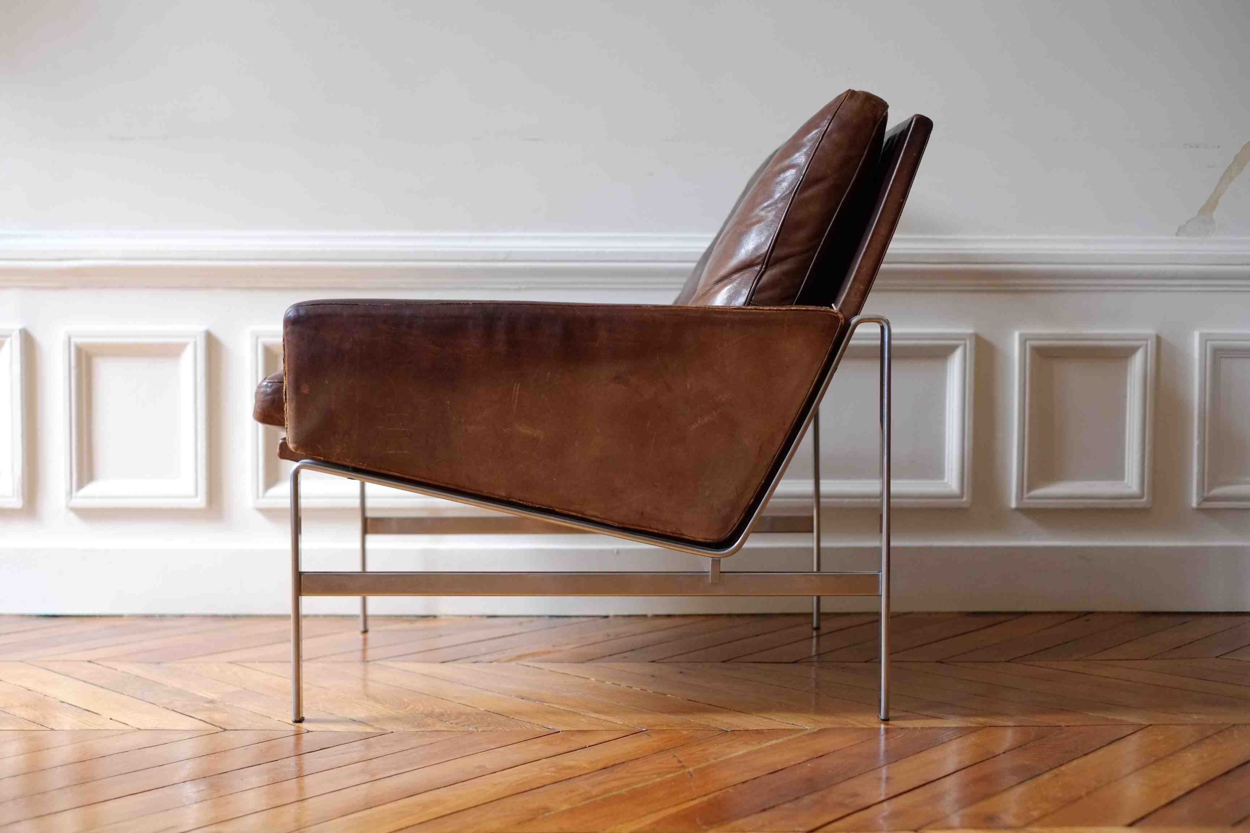 Mid-century modern leather armchair with slim metal legs and angled seat, placed on herringbone wood floors.