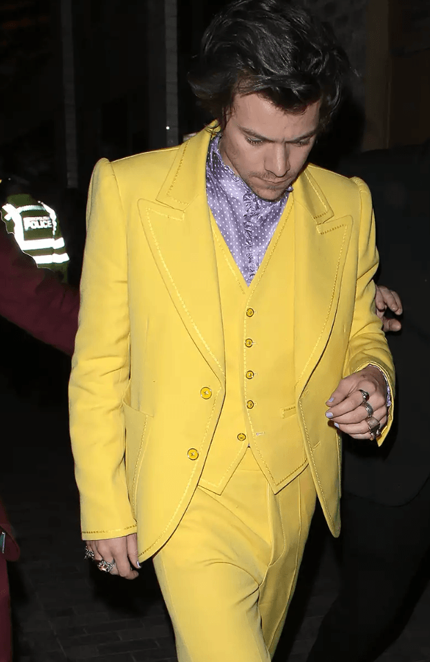 Harry Styles wearing a bright yellow three-piece suit with a lavender polka-dot shirt, walking at night with his head slightly bowed.